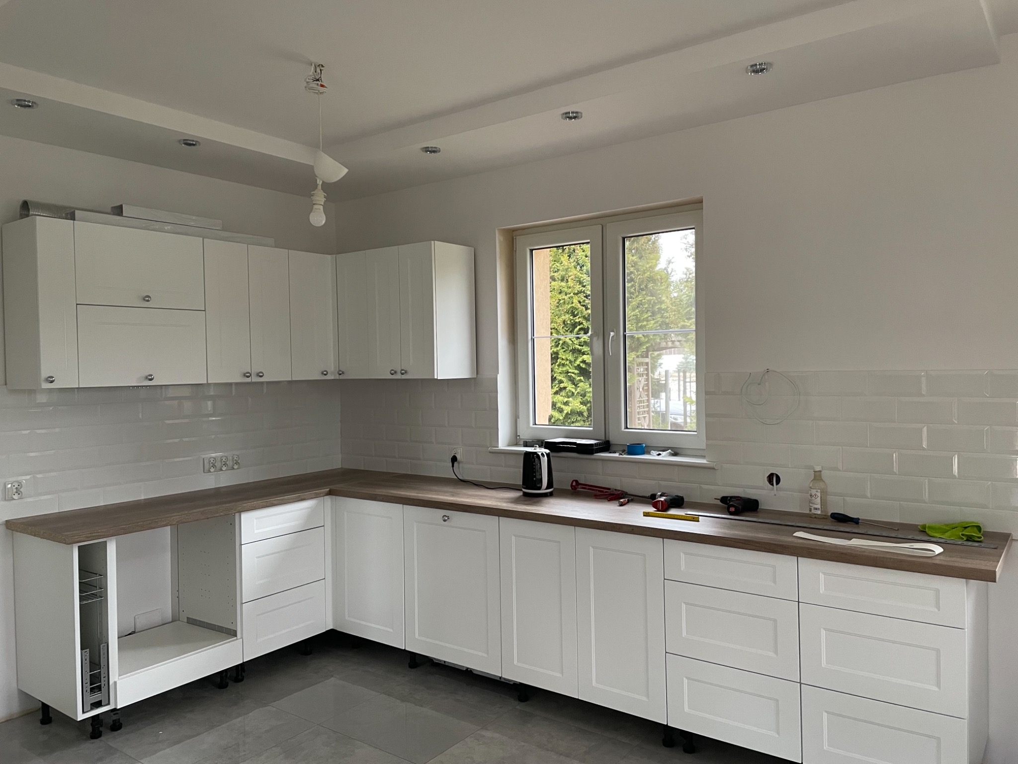 Wnętrze kuchni w trakcie montażu: białe szafki kuchenne z drewnianym blatem, białe płytki na ścianie, widoczne narzędzia i akcesoria montażowe, surowa żarówka zwisająca z sufitu.