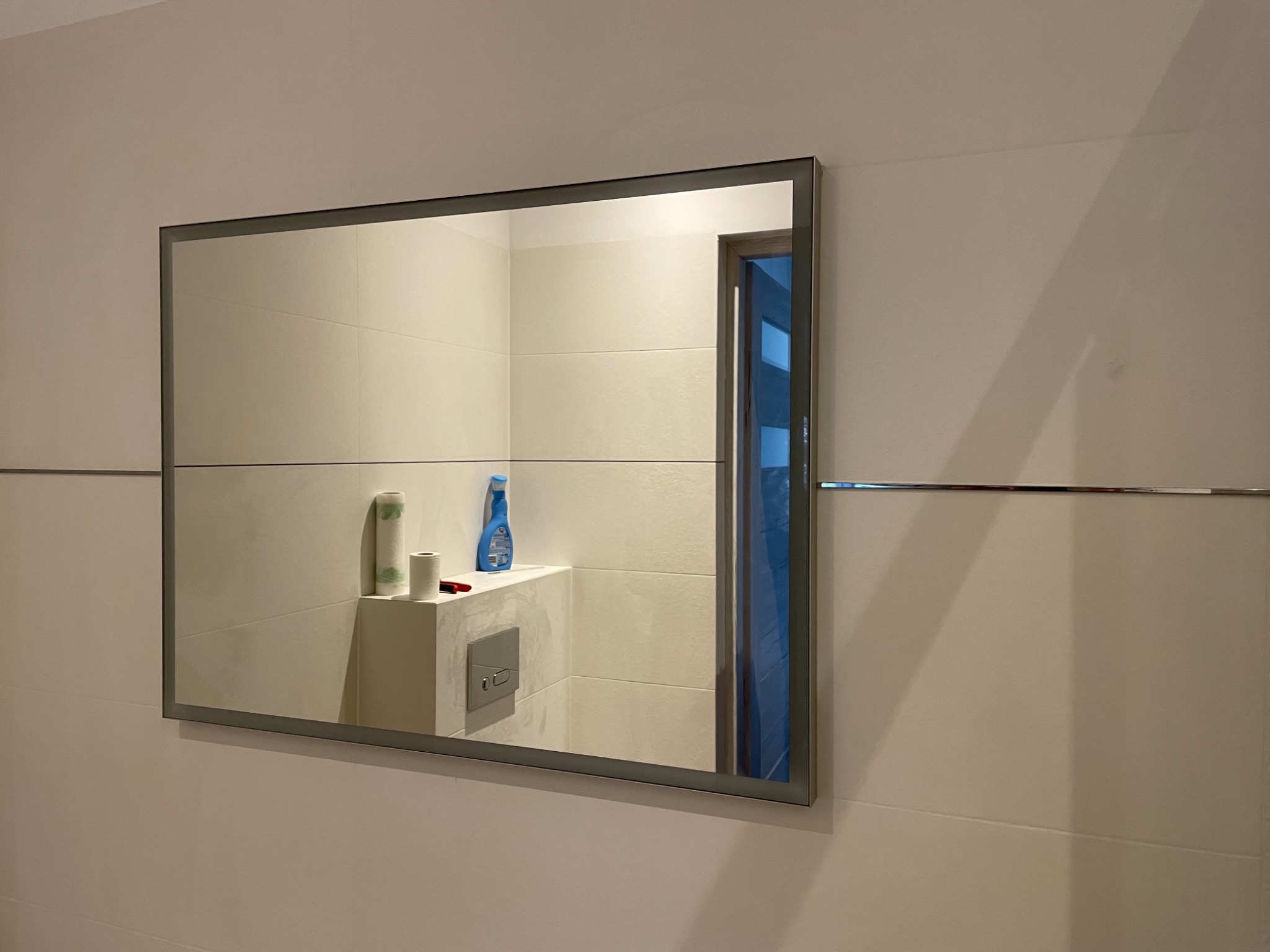 Prostokątne lustro w szarej ramie zawieszone na ścianie z płytek, odbijające fragment łazienki z toaletą, półką z pojemnikiem z płynem i rolką papieru toaletowego, oraz częściowo otwarte drzwi.