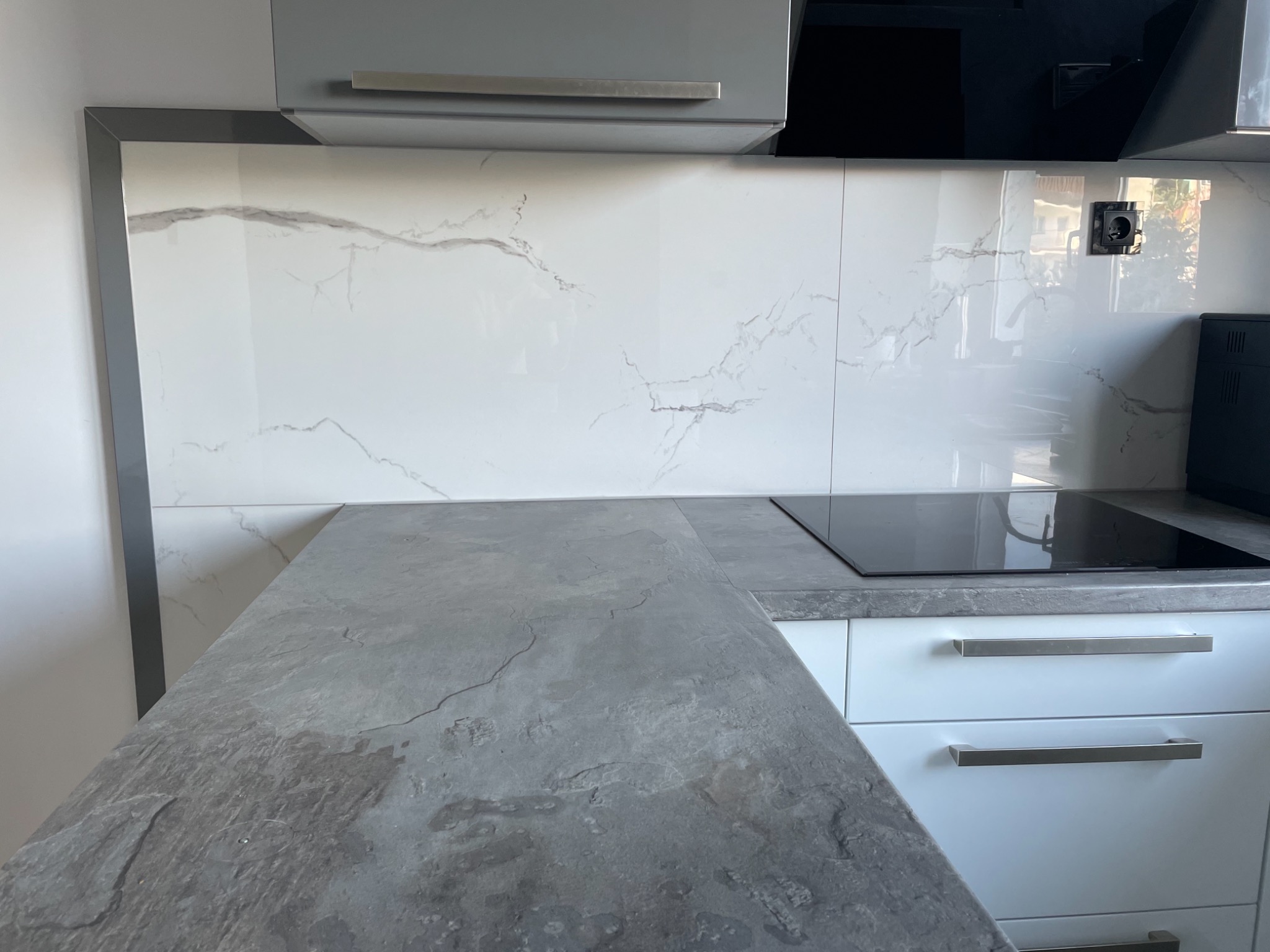 Fragment nowoczesnej kuchni z blatem imitującym kamień łupkowy, białe szafki z poziomymi, metalowymi uchwytami i płytki ścienne z marmurowym wzorem, widoczna płyta indukcyjna.