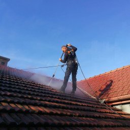 PRAMIX Mariusz Połeć - Pracownik w uprzęży, z bliska, czyści czerwoną dachówkę strumieniem wody pod ciśnieniem na tle błękitnego nieba.