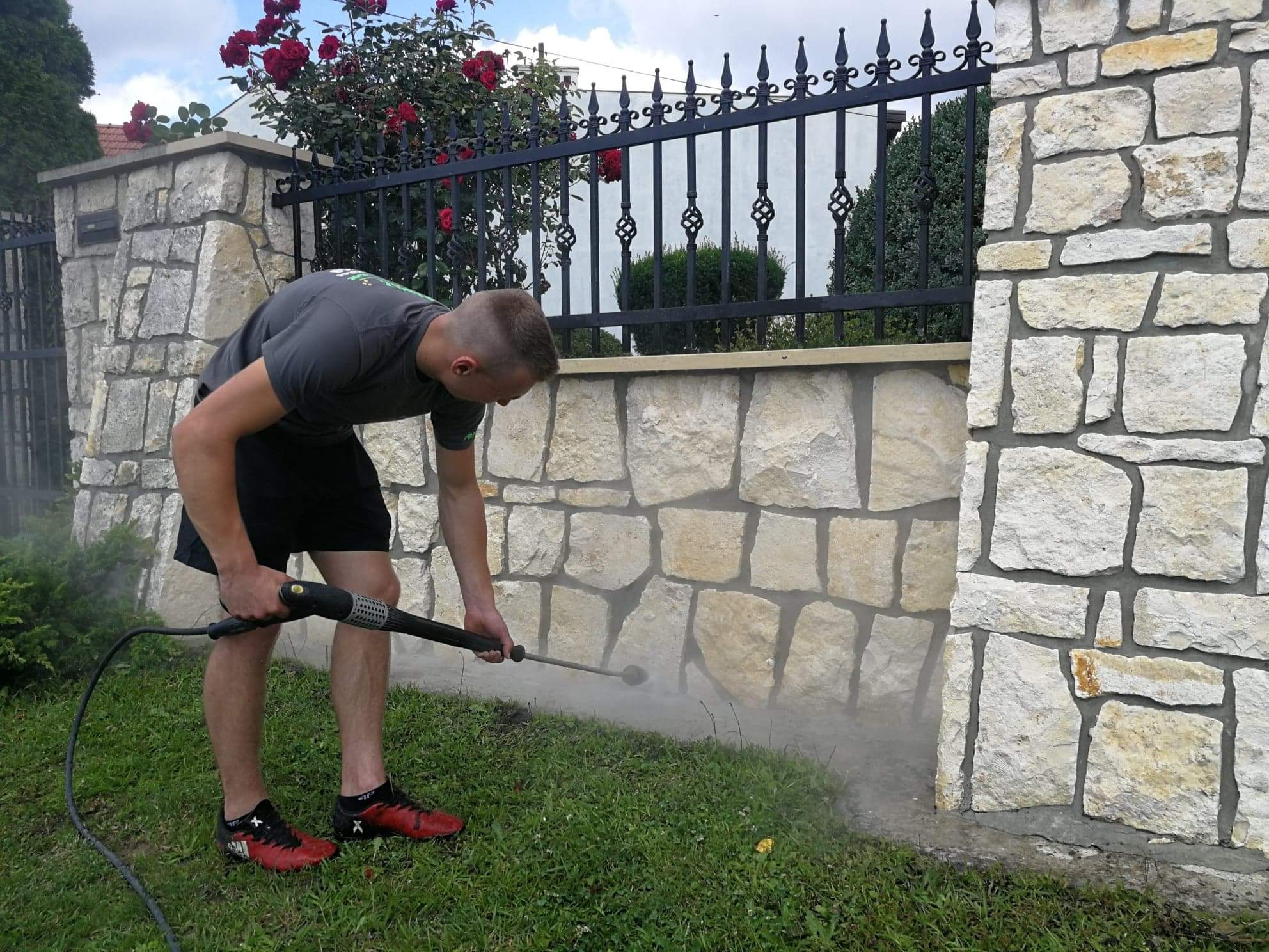 Mężczyzna w szarej koszulce i czarnych spodenkach czyści kamienny murek ogrodzeniowy za pomocą myjki ciśnieniowej, widoczne rozpryski wody i piana na trawie.