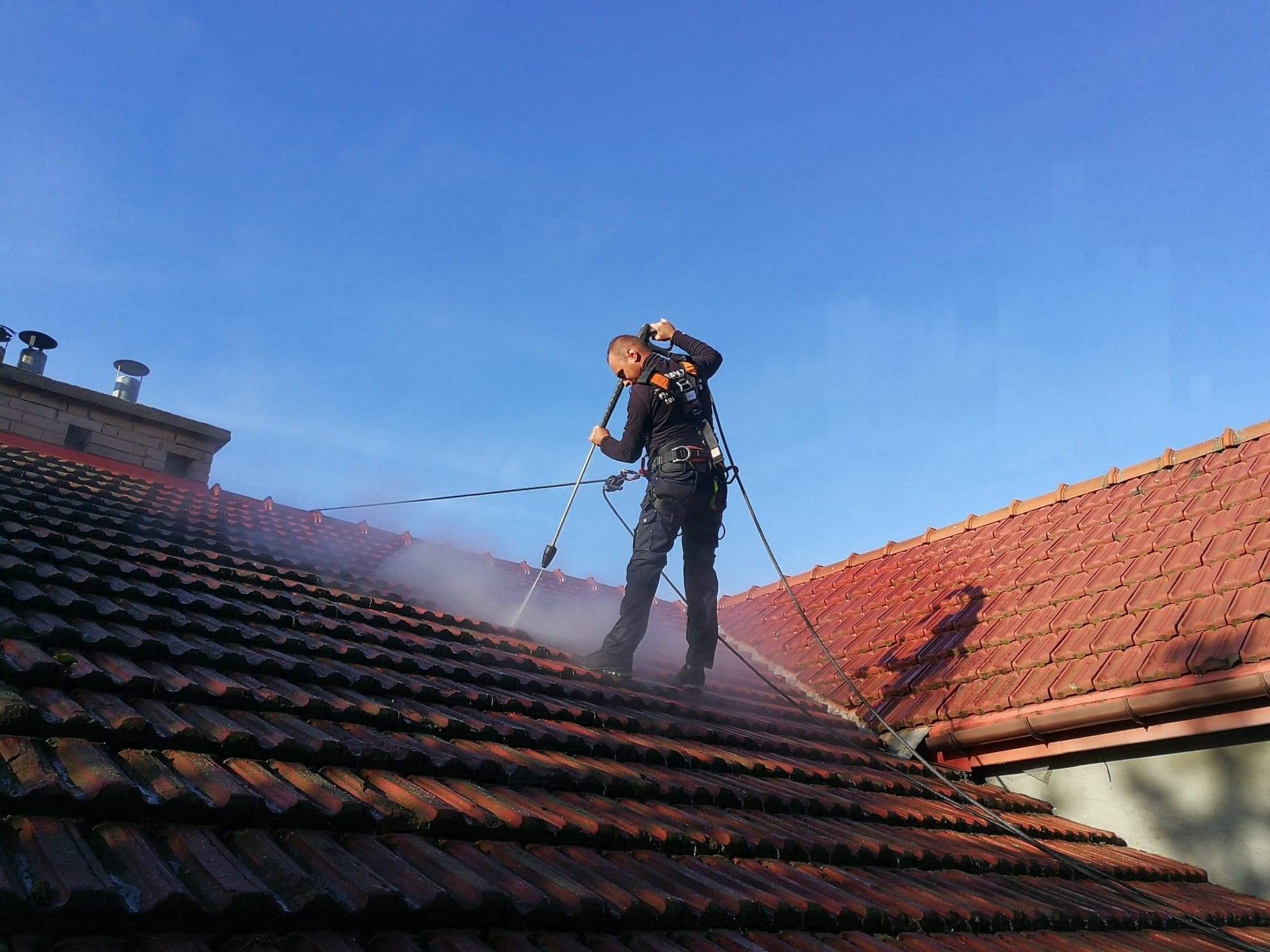 Pracownik w uprzęży, z bliska, czyści czerwoną dachówkę strumieniem wody pod ciśnieniem na tle błękitnego nieba.