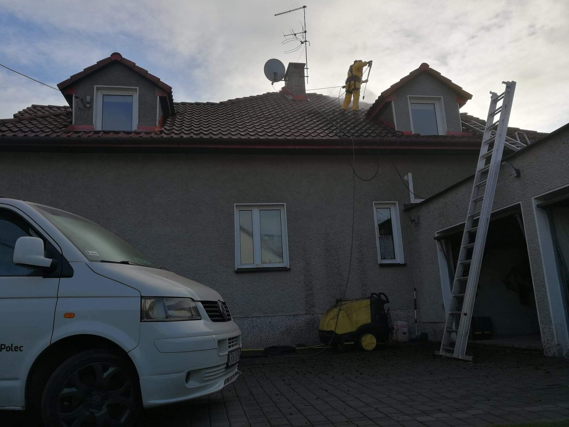 Pracownik w kombinezonie ochronnym czyści dach domu za pomocą myjki ciśnieniowej, widoczny samochód dostawczy i drabina oparta o budynek.
