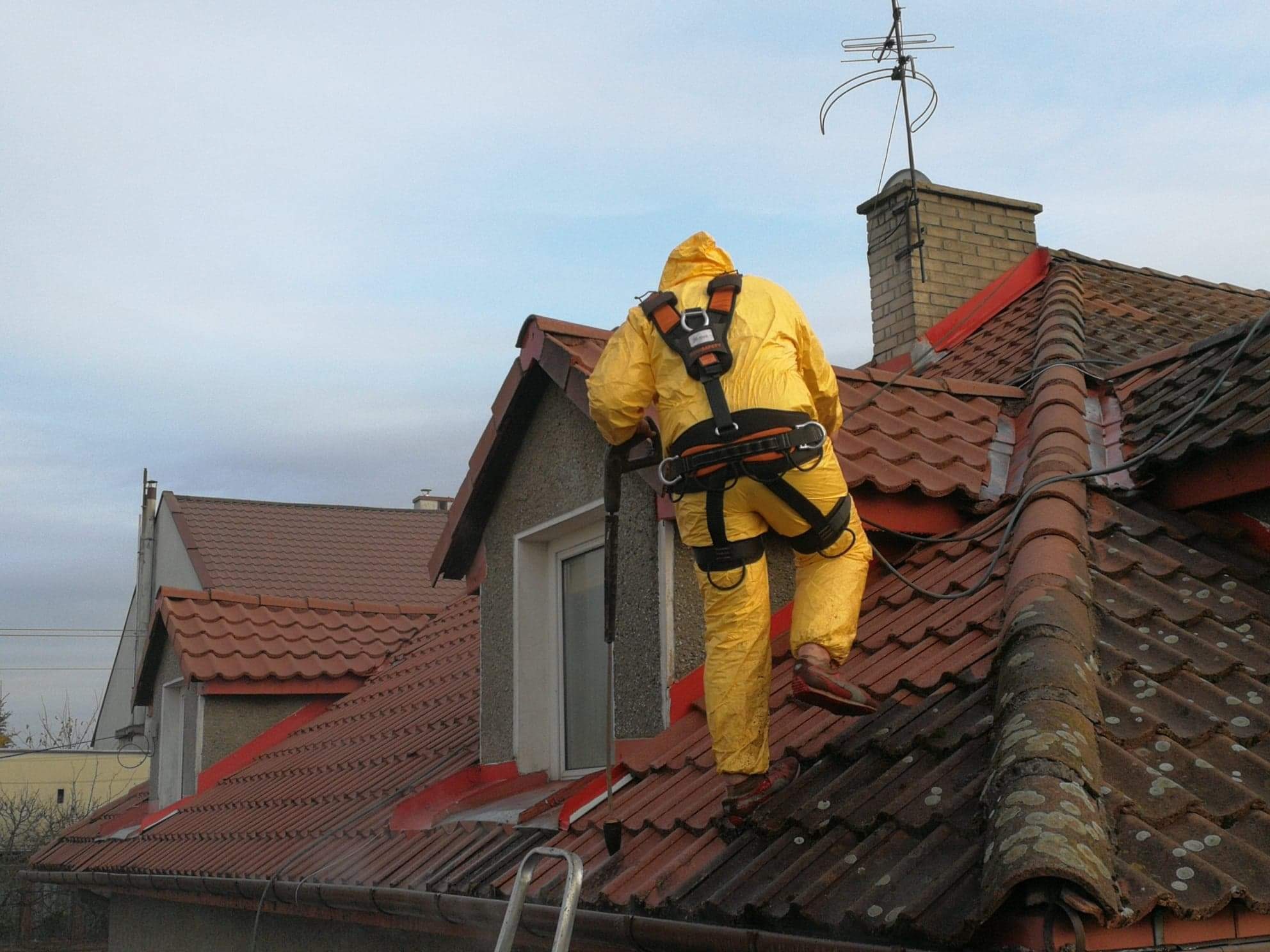 Pracownik w żółtym kombinezonie i uprzęży, z tyłu, czyści dachówkę za pomocą lancy ciśnieniowej, widoczny mech na części dachu.