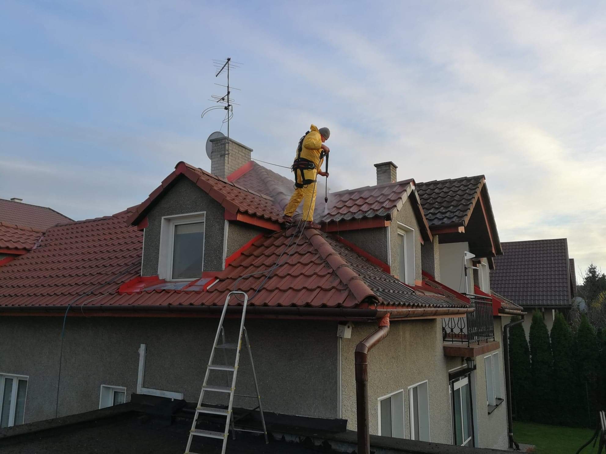 Pracownik w żółtym kombinezonie i uprzęży, stojący na czerwonym dachu domu i spryskujący go wodą pod ciśnieniem, widoczna drabina oparta o budynek.