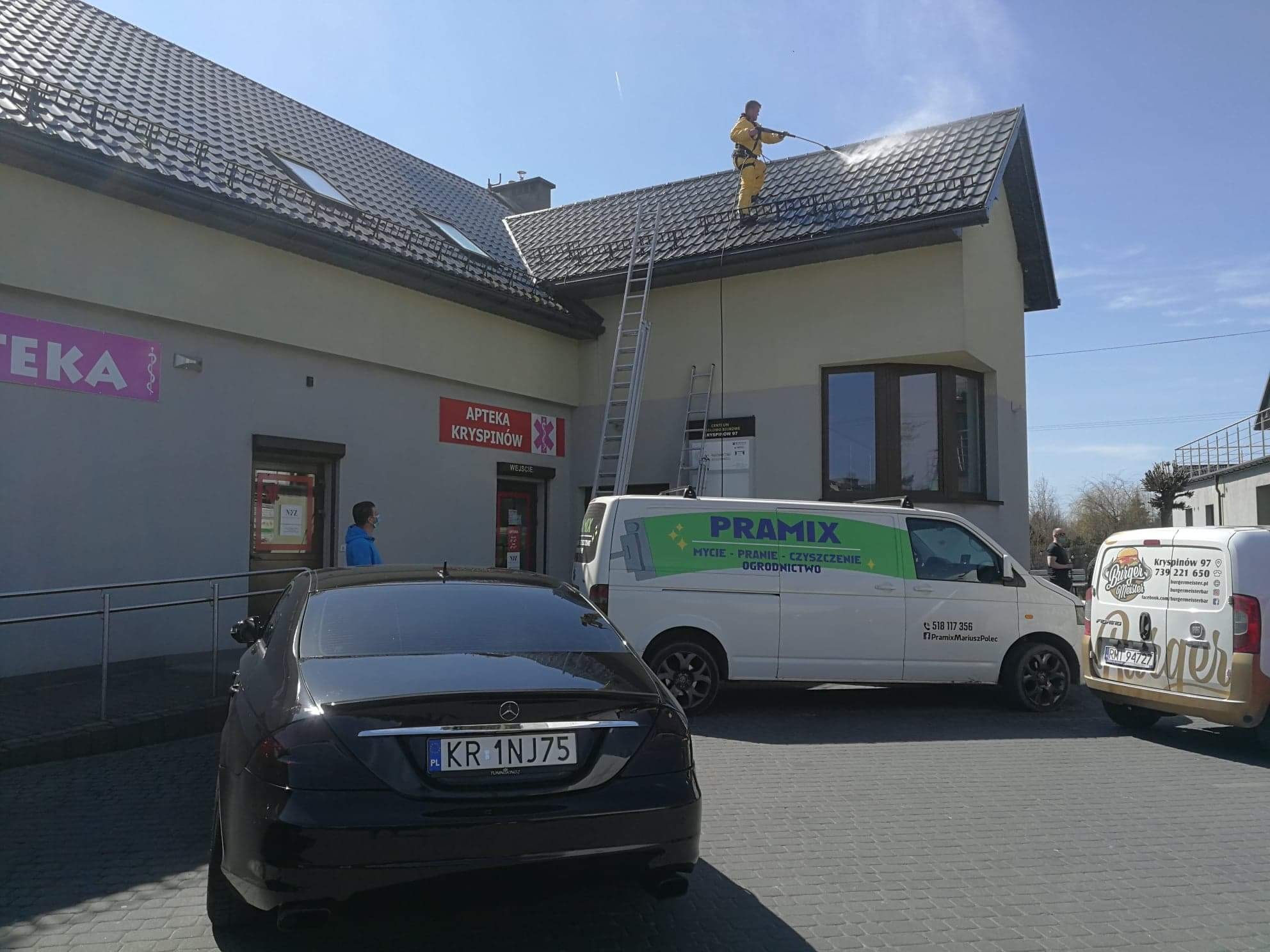 Pracownik w żółtym kombinezonie myje dach budynku z szarej dachówki za pomocą myjki ciśnieniowej, widoczna drabina, zaparkowany czarny Mercedes i biały bus z logo firmy świadczącej usługi czyszczące.