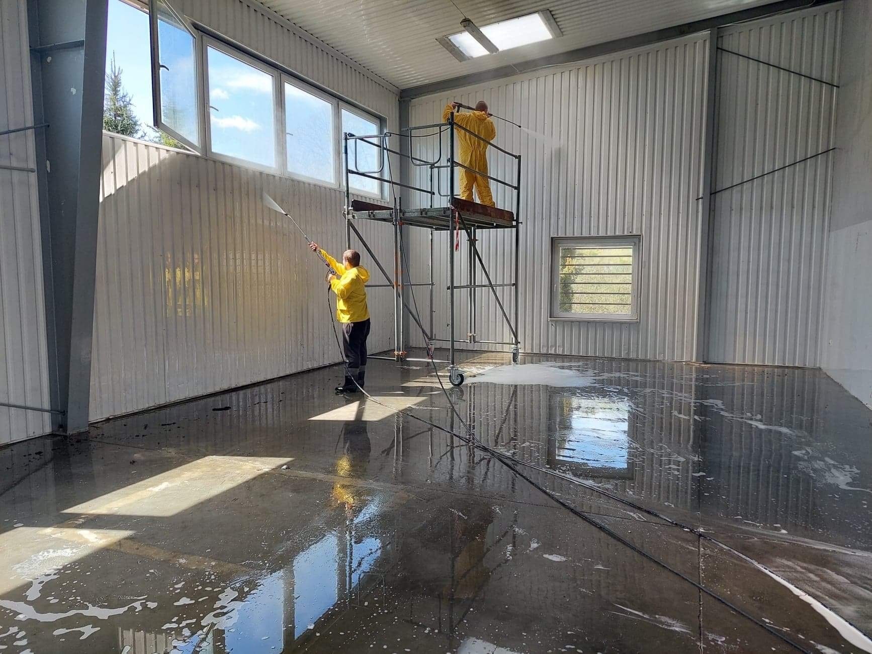 Dwóch pracowników w kombinezonach ochronnych myje ciśnieniowo wnętrze hali produkcyjnej, jeden z nich stoi na rusztowaniu na kółkach.