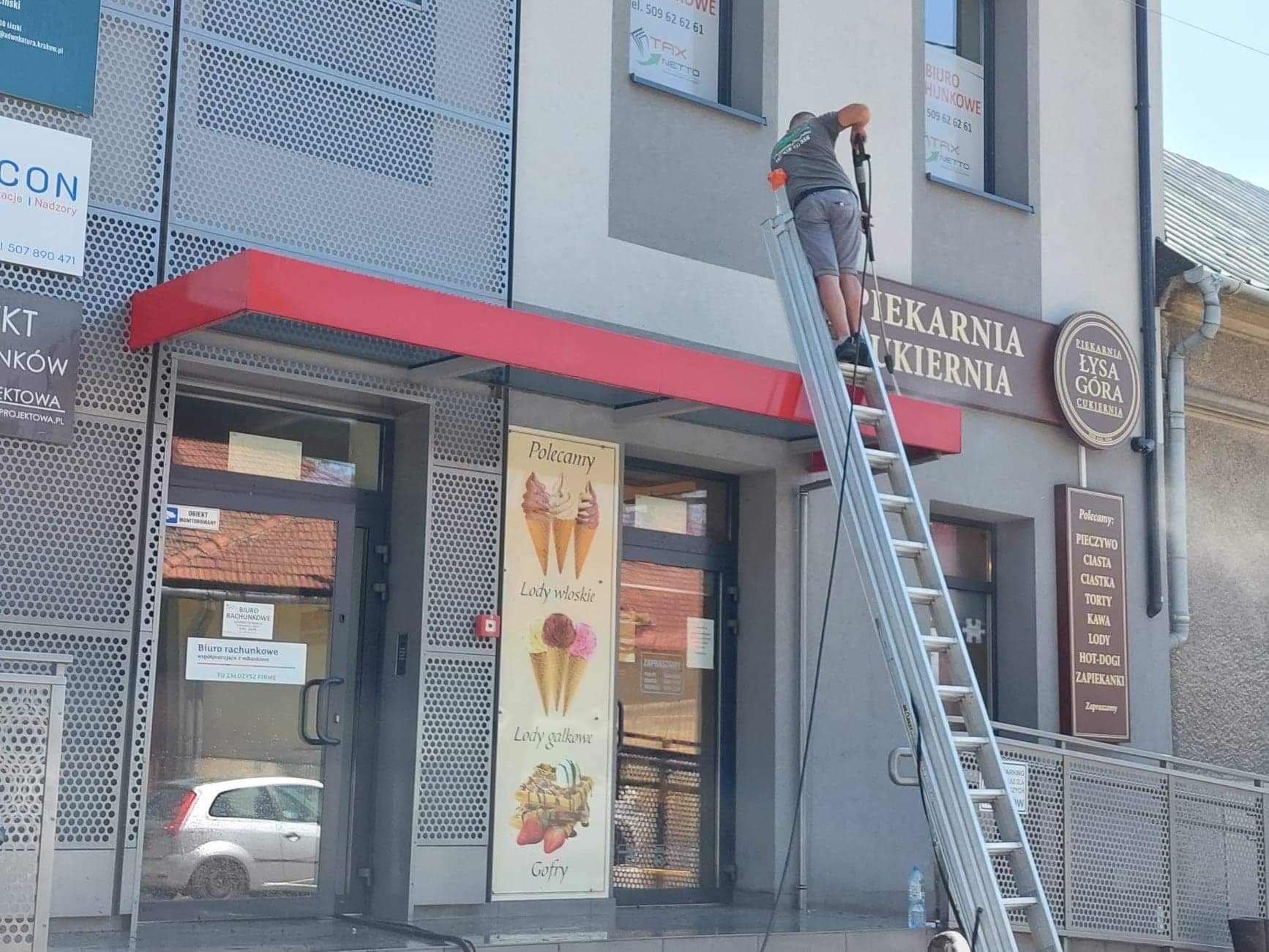 Mężczyzna na drabinie, z narzędziem w ręku, prawdopodobnie czyszczący elewację budynku piekarni z szyldem 'Piekarnia Łysa Góra Cukiernia', widoczny fragment perforowanej elewacji i plakaty z ofertą...