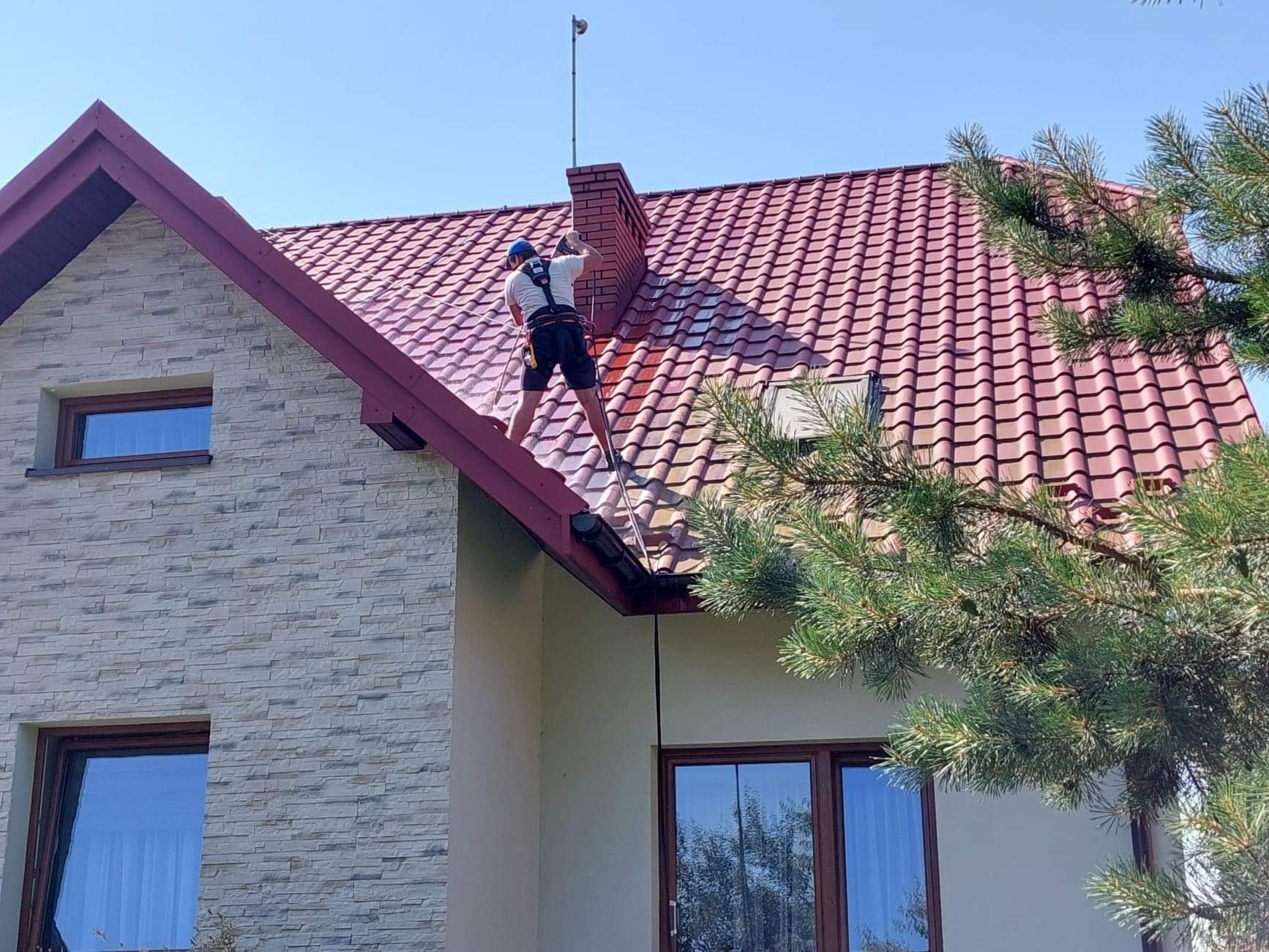Pracownik w uprzęży na dachu domu z czerwonej dachówki, czyści dach lancą ciśnieniową. Widoczny komin i fragment elewacji budynku wykończonej kamieniem dekoracyjnym.