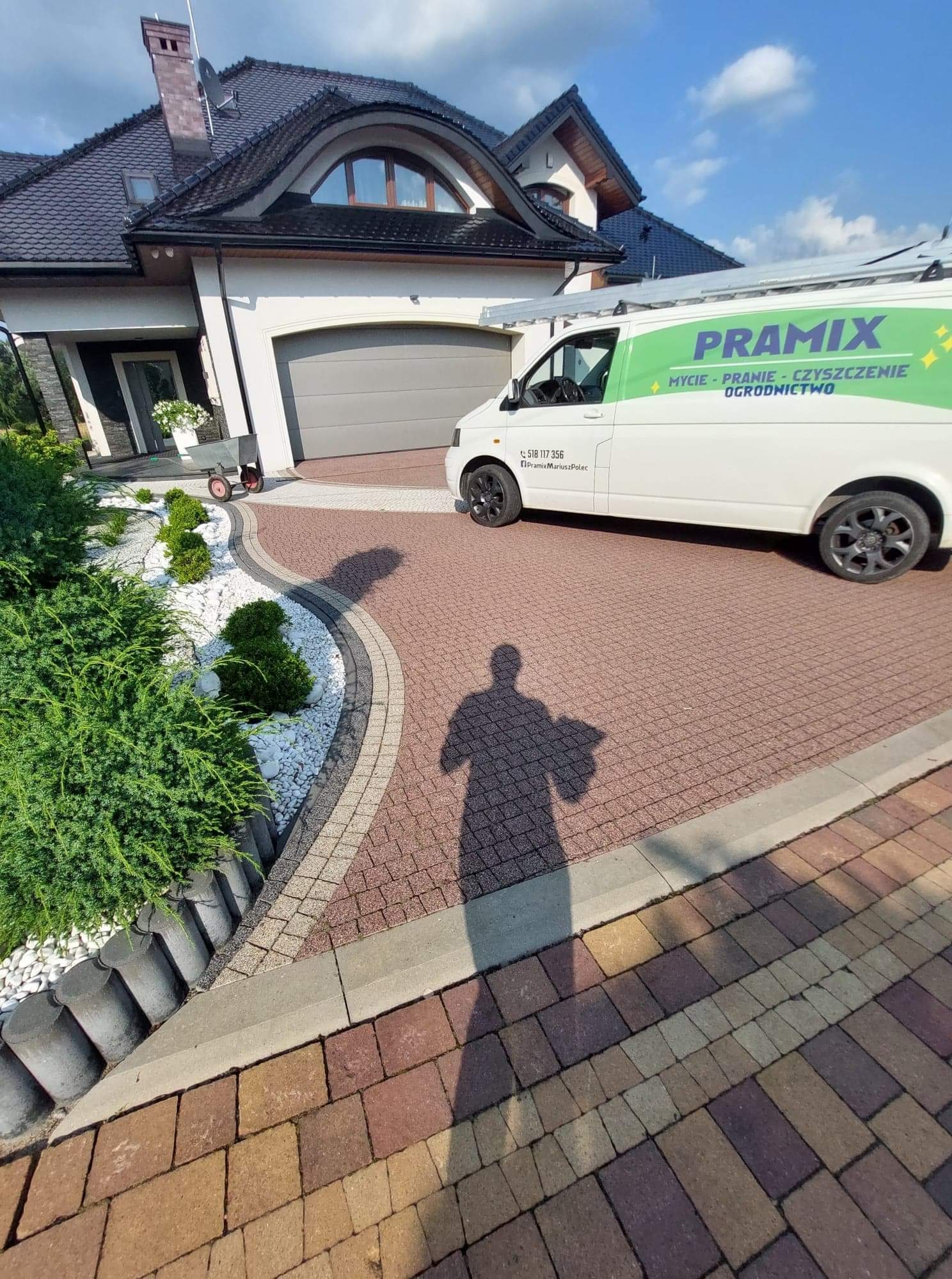 Nowoczesny dom jednorodzinny z zadbanym ogrodem, podjazd z kostki brukowej, biały bus firmy z logo i hasłem 'Mycie - Pranie - Czyszczenie - Ogrodnictwo', cień fotografa na pierwszym planie.