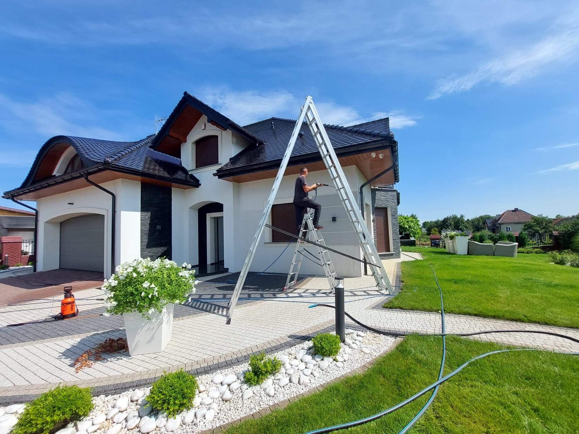 Mężczyzna w czarnym ubraniu myje elewację domu ciśnieniowo, stojąc na rozstawionej aluminiowej drabinie na tle błękitnego nieba i zadbanego ogrodu.