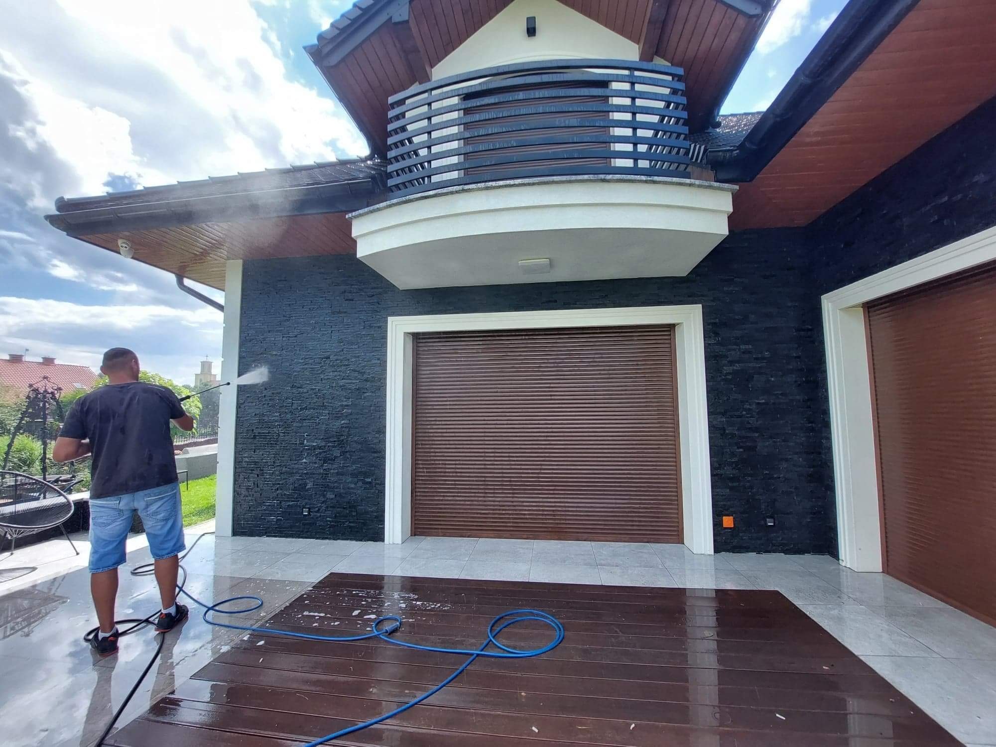 Mężczyzna czyści elewację domu z czarnej cegły za pomocą myjki ciśnieniowej, widoczny rozprysk wody i mokra powierzchnia tarasu.