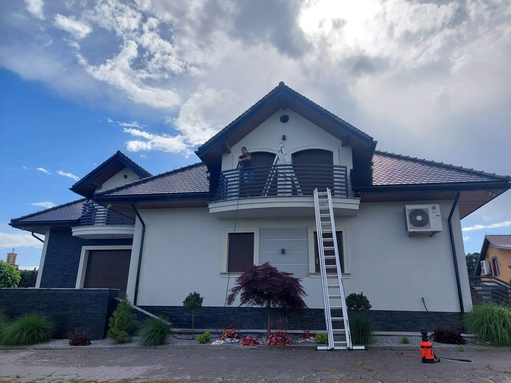 Pracownik na balkonie domu z drabiny czyści elewację za pomocą lancy ciśnieniowej; widoczny spryskiwacz ciśnieniowy na chodniku.