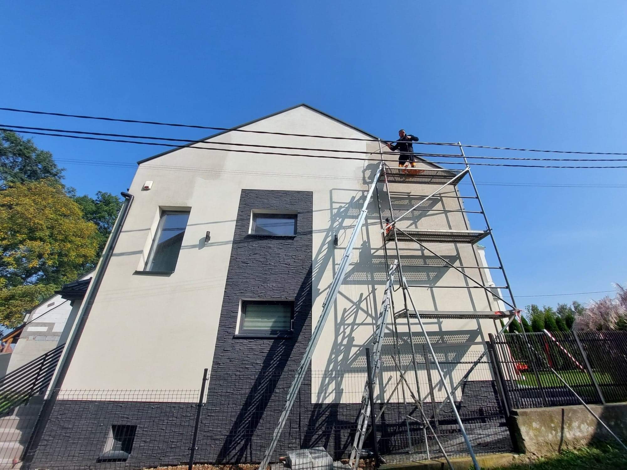 Pracownik na rusztowaniu, malujący elewację dwukondygnacyjnego domu z fragmentem elewacji wykończonym ciemnym kamieniem dekoracyjnym, widoczne przewody elektryczne na tle błękitnego nieba.