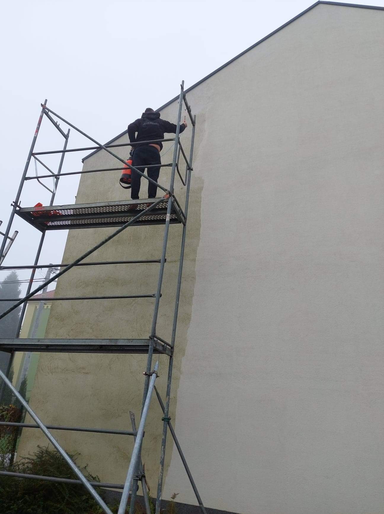 Pracownik na rusztowaniu, z pomarańczowym pojemnikiem, aplikuje preparat na elewację budynku, widoczny fragment odświeżonej powierzchni.