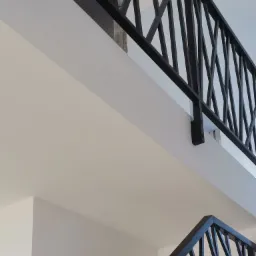 Wnętrze z czarną, metalową balustradą o geometrycznym wzorze, widok z dołu na dwa poziomy, białe ściany i sufit.