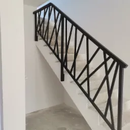 Nowoczesna, czarna barierka schodowa z geometrycznym wzorem, zamontowana na betonowych schodach w surowym stanie.