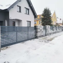Nowoczesna, pozioma brama przesuwna w kolorze antracytowym z murowanymi słupkami z szarej kostki, przed domem z numerem 11, zimowy krajobraz pokryty śniegiem.