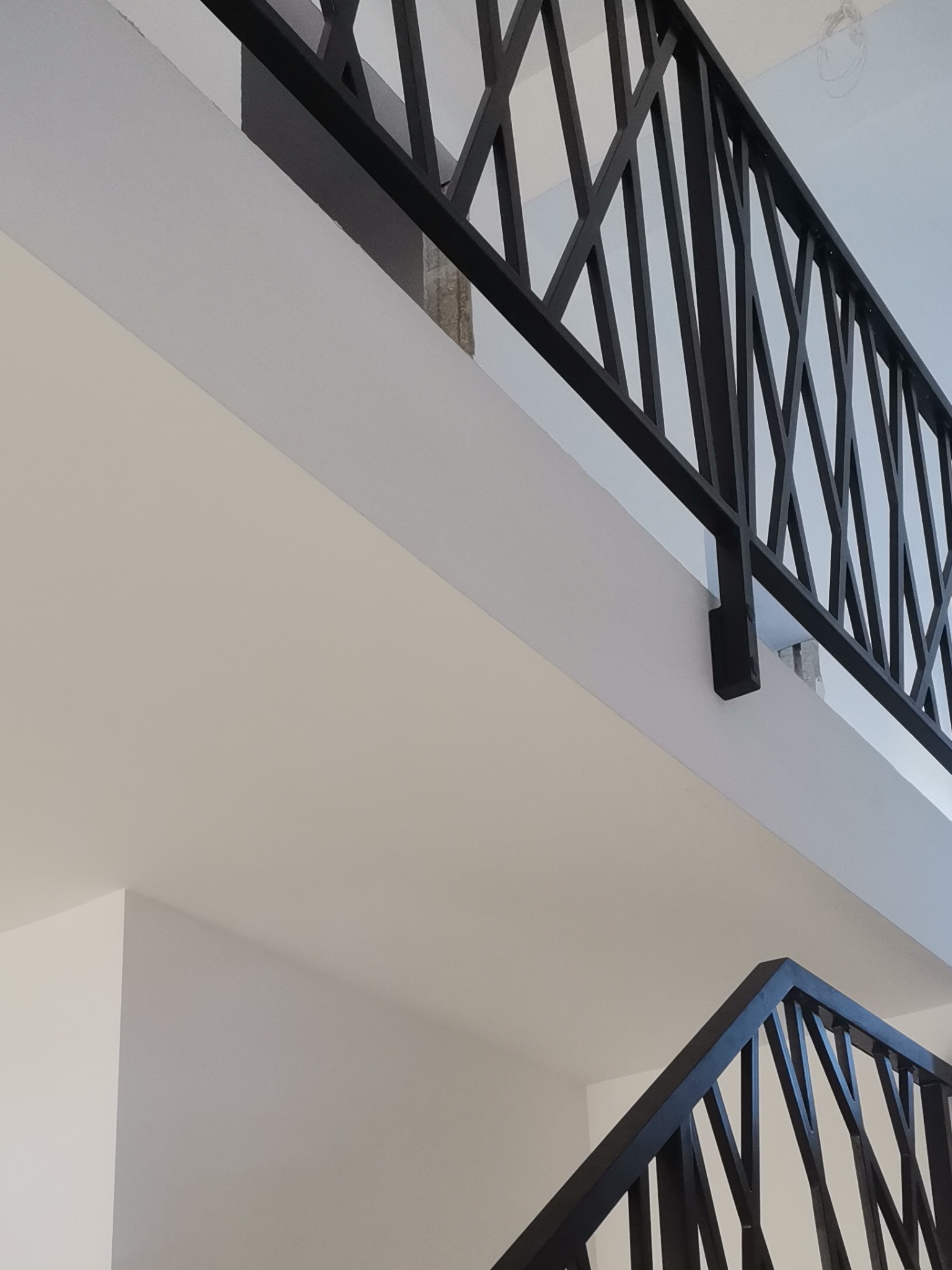 Wnętrze z czarną, metalową balustradą o geometrycznym wzorze, widok z dołu na dwa poziomy, białe ściany i sufit.