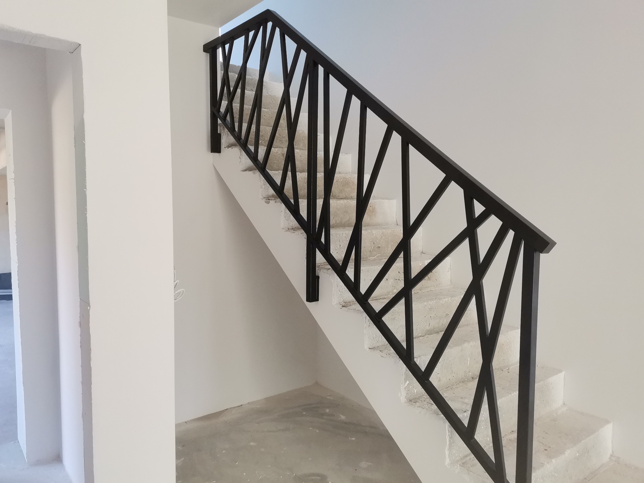 Nowoczesna, czarna barierka schodowa z geometrycznym wzorem, zamontowana na betonowych schodach w surowym stanie.