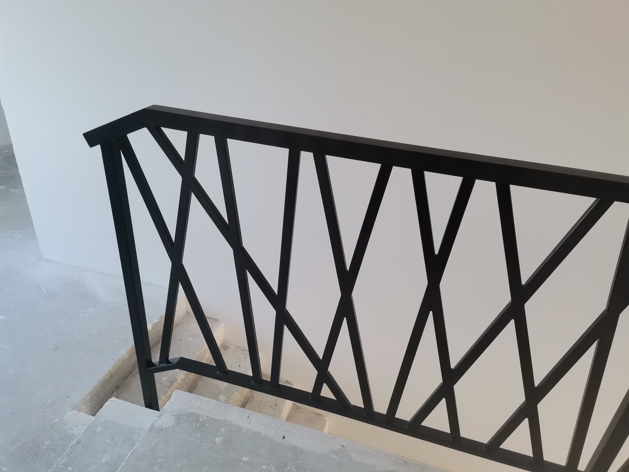 Nowoczesna, czarna balustrada schodowa z geometrycznym wzorem ukośnych linii, zamontowana na surowych, betonowych schodach w jasnym wnętrzu w trakcie wykończenia.