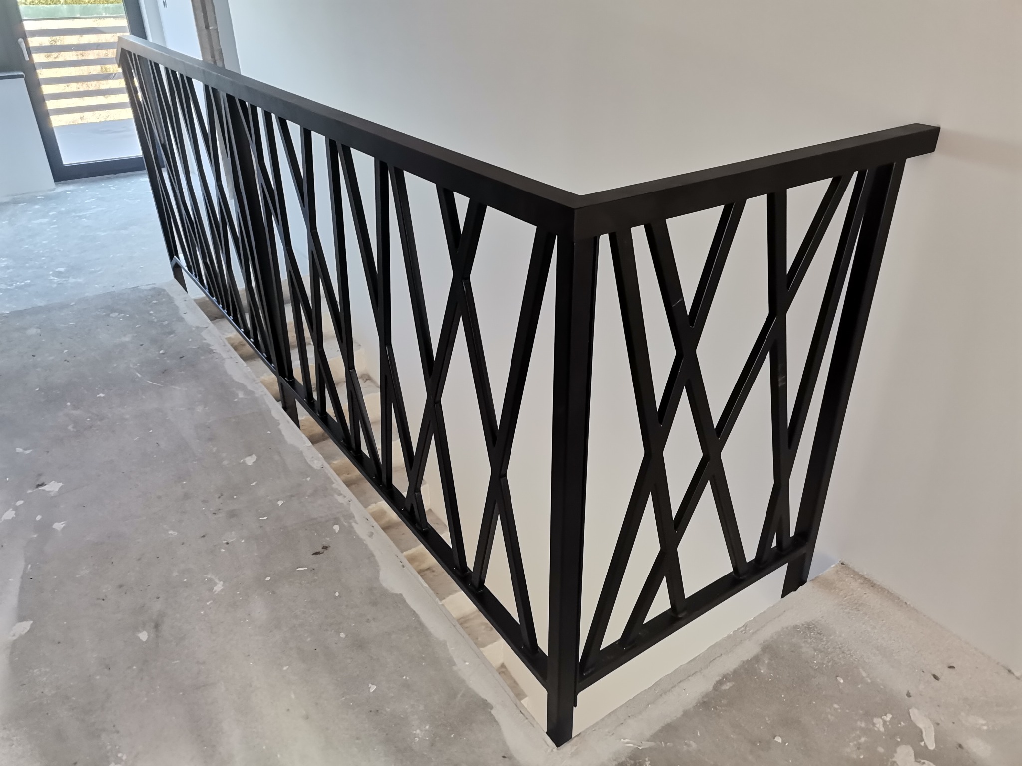 Czarna metalowa balustrada schodowa o geometrycznym wzorze krzyżujących się linii, zamontowana w jasnym wnętrzu podczas prac wykończeniowych, widoczny surowy beton na podłodze.