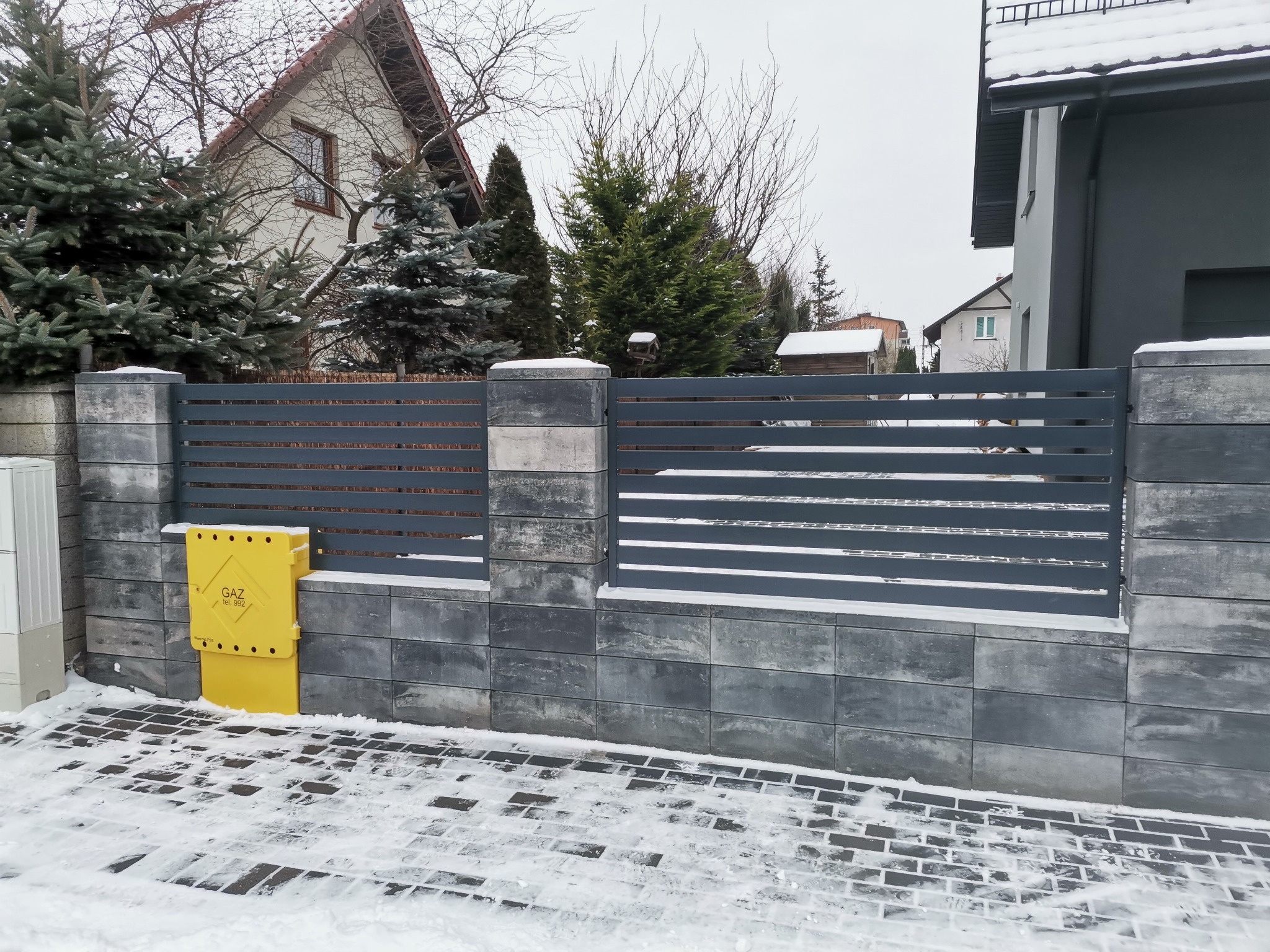 Nowoczesne ogrodzenie panelowe w kolorze antracytowym, zbudowane z betonowych bloczków, z zaśnieżonym chodnikiem i żółtą skrzynką gazową, na tle zimowego krajobrazu.