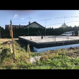 Przygotowany fundament z izolacją pionową, widoczne wystające pręty zbrojeniowe, na działce zarośniętej trawą, w tle dom jednorodzinny i zaparkowany samochód dostawczy.