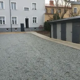 Wykończony teren posesji z wysypanym szarym grysem, widok na budynek mieszkalny z szarą elewacją i rząd garaży z ciemnymi drzwiami.
