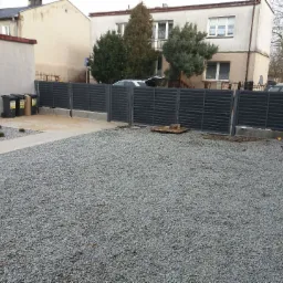 Nowoczesne, poziome ogrodzenie panelowe w kolorze antracytowym, oddzielające posesję wysypaną szarym żwirem od sąsiednich budynków; widoczne śmietniki i fragmenty budynków w tle.