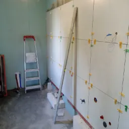 Ściana w trakcie układania białych płytek z użyciem poziomicy, sznurków i klipsów dystansowych; widoczne otwory na instalacje, drabina i bloczki betonowe na podłodze.