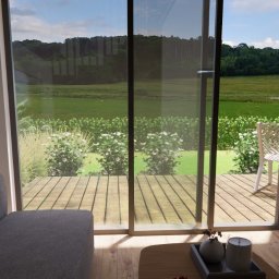 OGRODOWSKA Katarzyna Prokopiuk - Wnętrze domu z widokiem na ogród przez przesuwne szklane drzwi, z widoczną częścią szarej sofy i minimalistycznym stolikiem kawowym z dekoracjami, oraz wysokimi stołkami barowymi przy drewnianym...