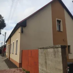 Odnowiona elewacja dwukondygnacyjnego domu z brązowym akcentem, widoczna z poziomu chodnika, z zamontowaną anteną satelitarną i pomalowanym na pomarańczowo ogrodzeniem w tle.