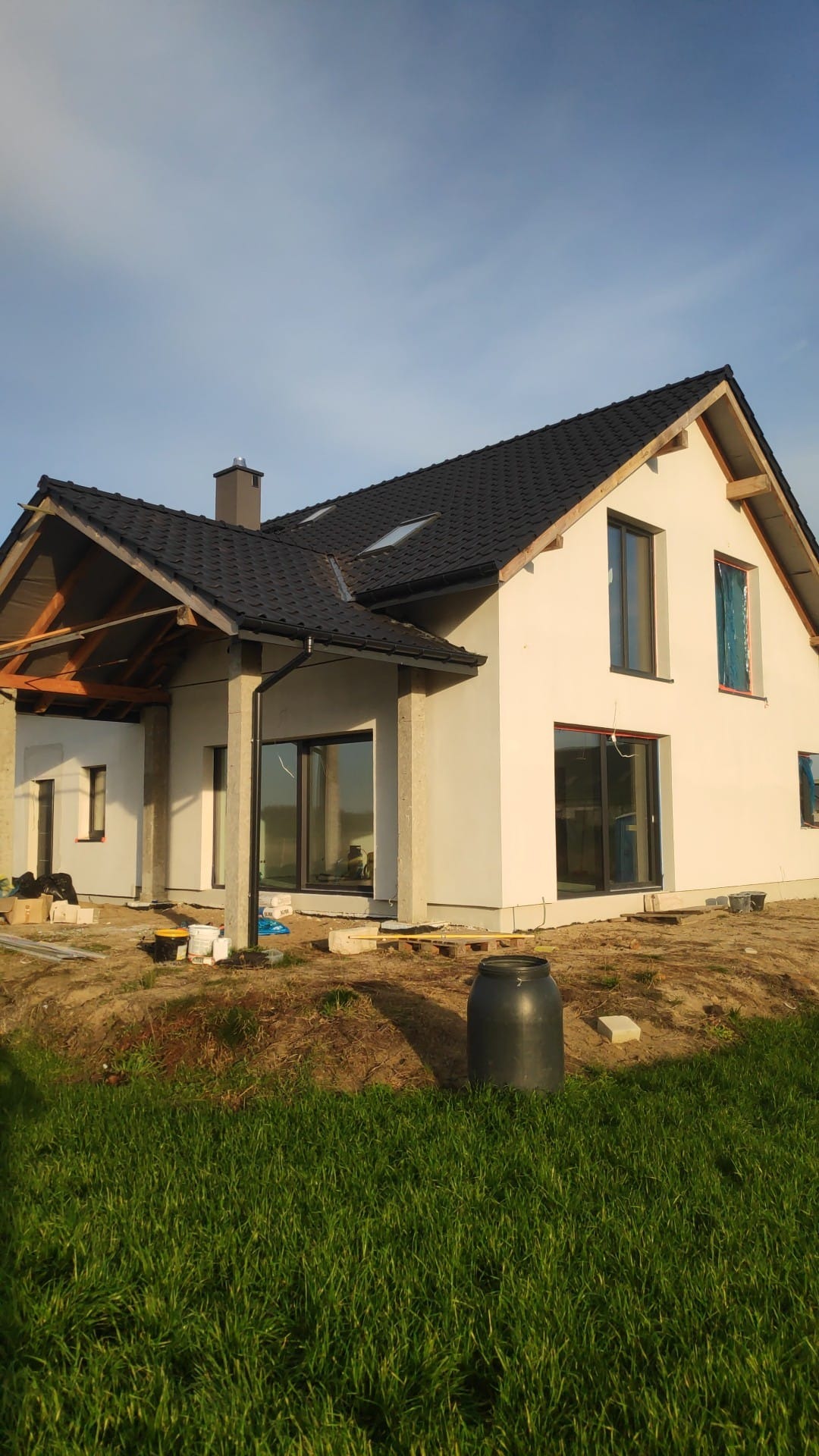 Niedokończony dom jednorodzinny z czarnym dachem i oknami, częściowo otynkowany, z widocznymi elementami konstrukcyjnymi werandy i materiałami budowlanymi na placu budowy.
