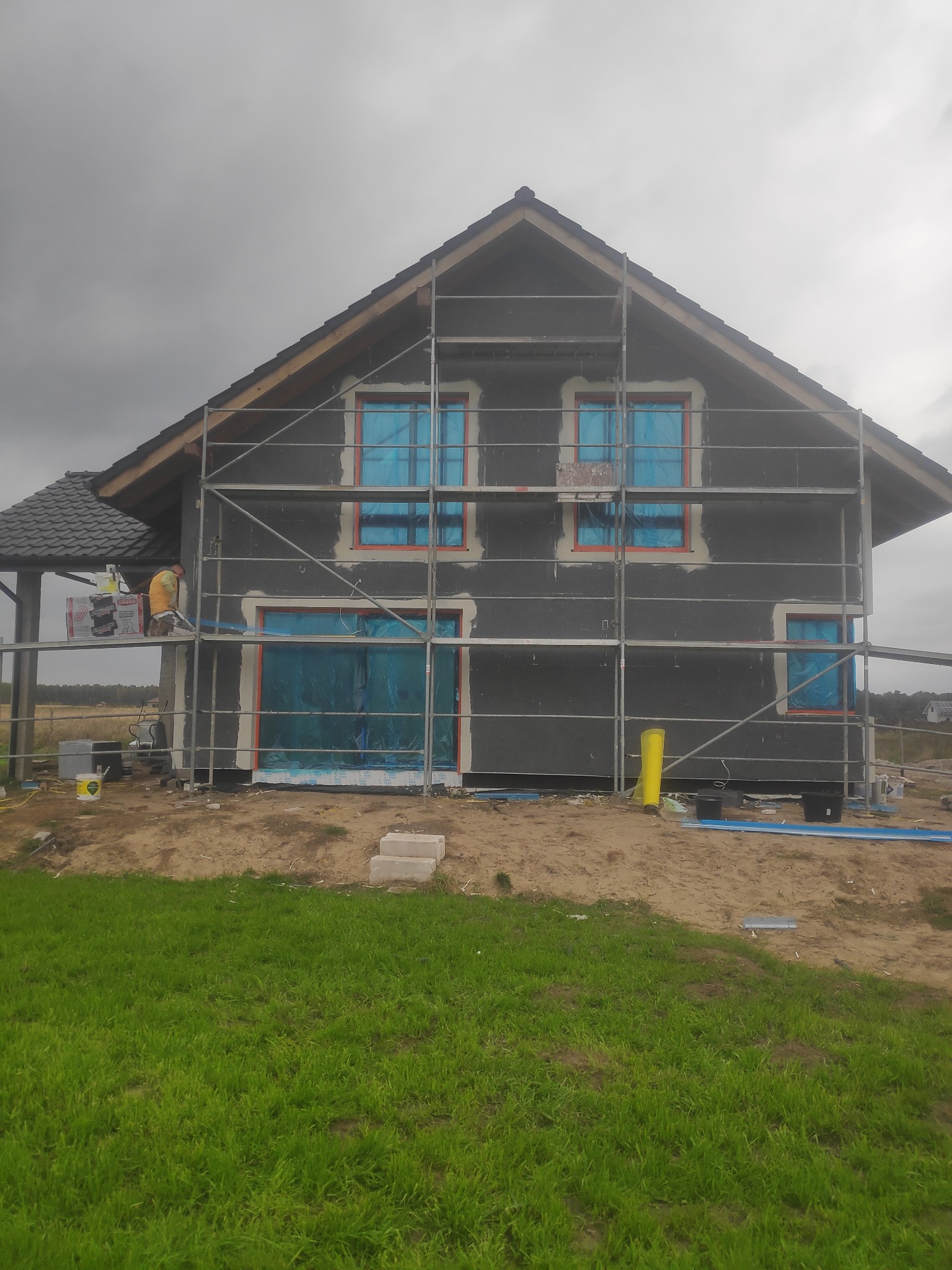 Dom w trakcie ocieplania elewacji, okna zabezpieczone folią, widoczne rusztowania i pracownik w odblaskowej kamizelce, przygotowujący materiały izolacyjne.