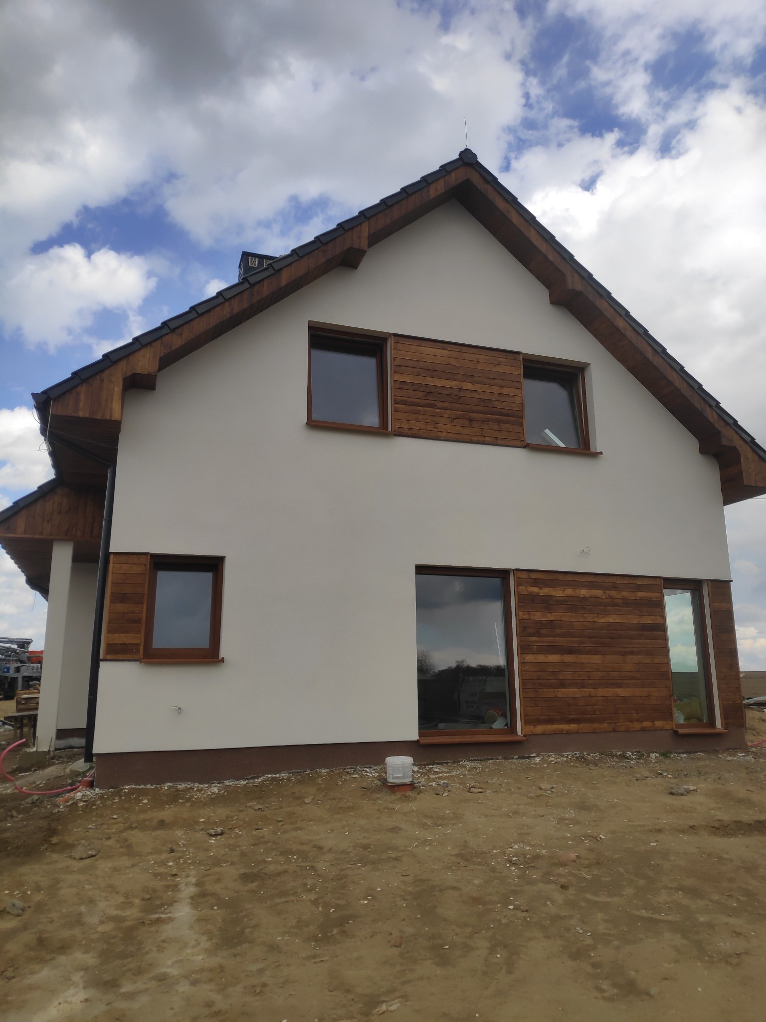 Nowoczesny dom jednorodzinny z białą elewacją i drewnianymi akcentami wokół okien, widoczne elementy wykończeniowe i teren wokół budynku w trakcie prac.