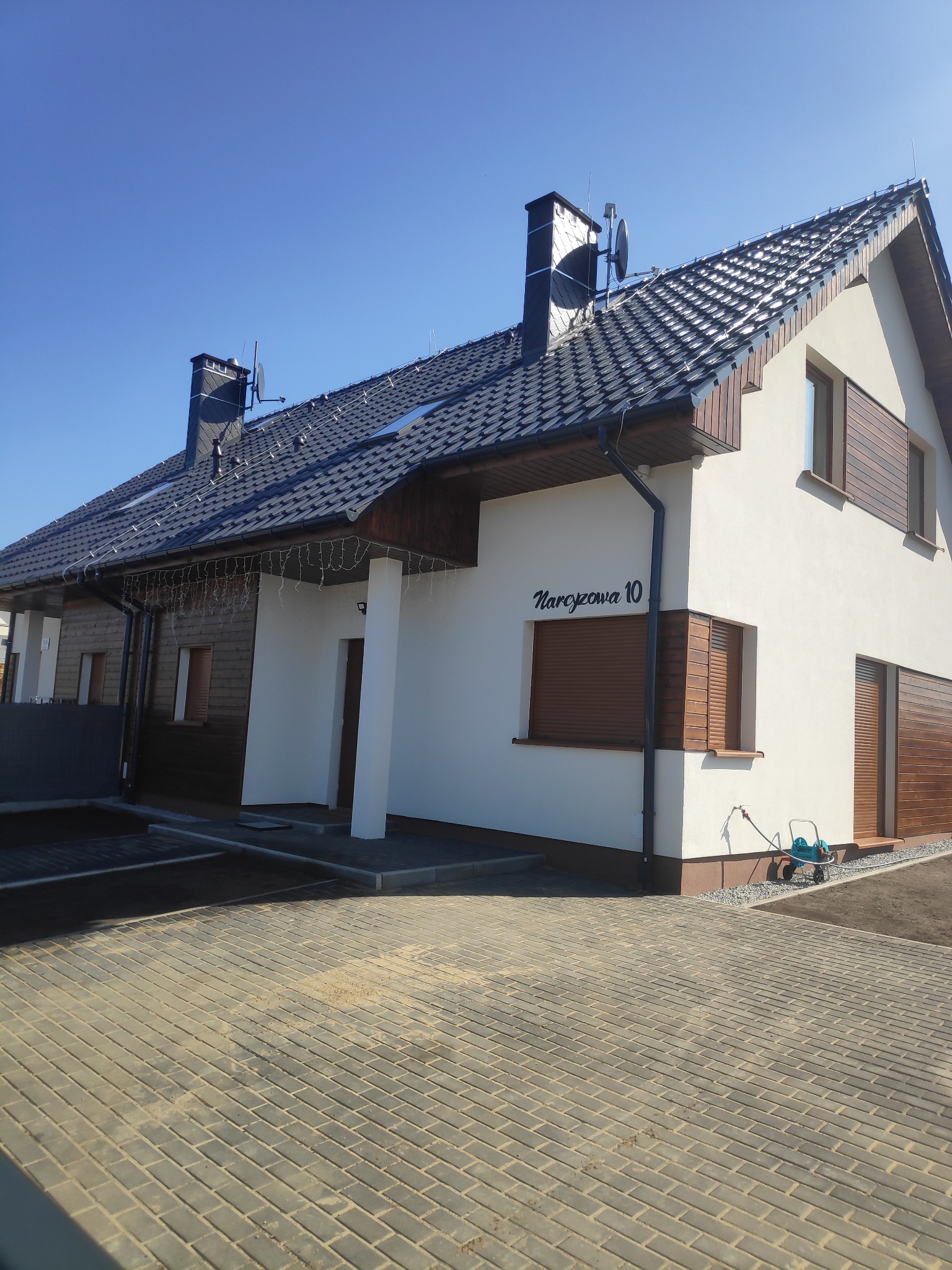 Nowoczesny dom jednorodzinny z białą elewacją, drewnianymi akcentami i ciemną dachówką, adres 'Narcyzowa 10' widoczny na ścianie.