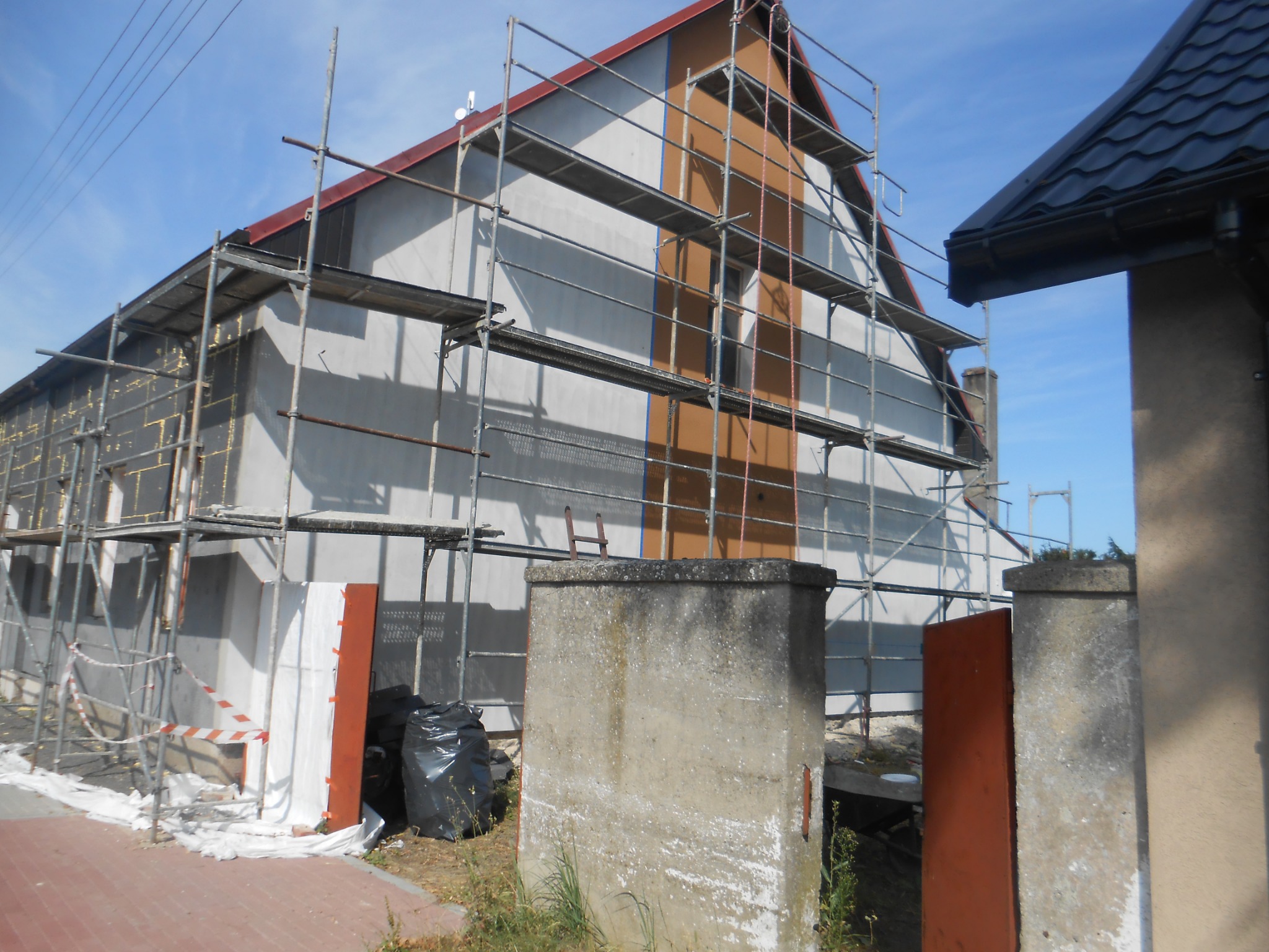 Dom jednorodzinny w trakcie ocieplania, widoczne rusztowania, częściowo ocieplona ściana z żółtą izolacją, fragment ściany pomalowany na brązowo, białe płyty styropianowe opierają się o ścianę.