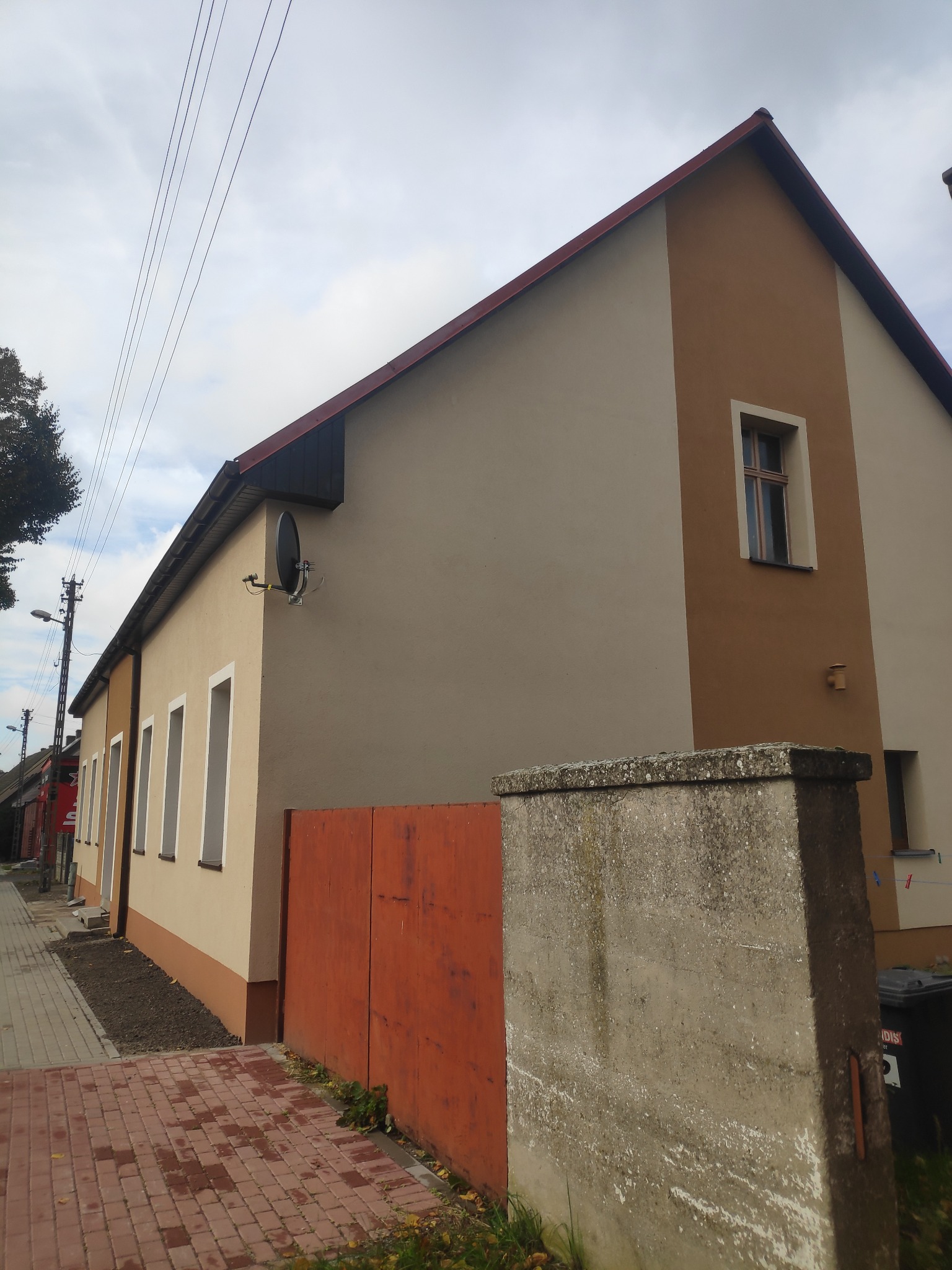 Odnowiona elewacja dwukondygnacyjnego domu z brązowym akcentem, widoczna z poziomu chodnika, z zamontowaną anteną satelitarną i pomalowanym na pomarańczowo ogrodzeniem w tle.