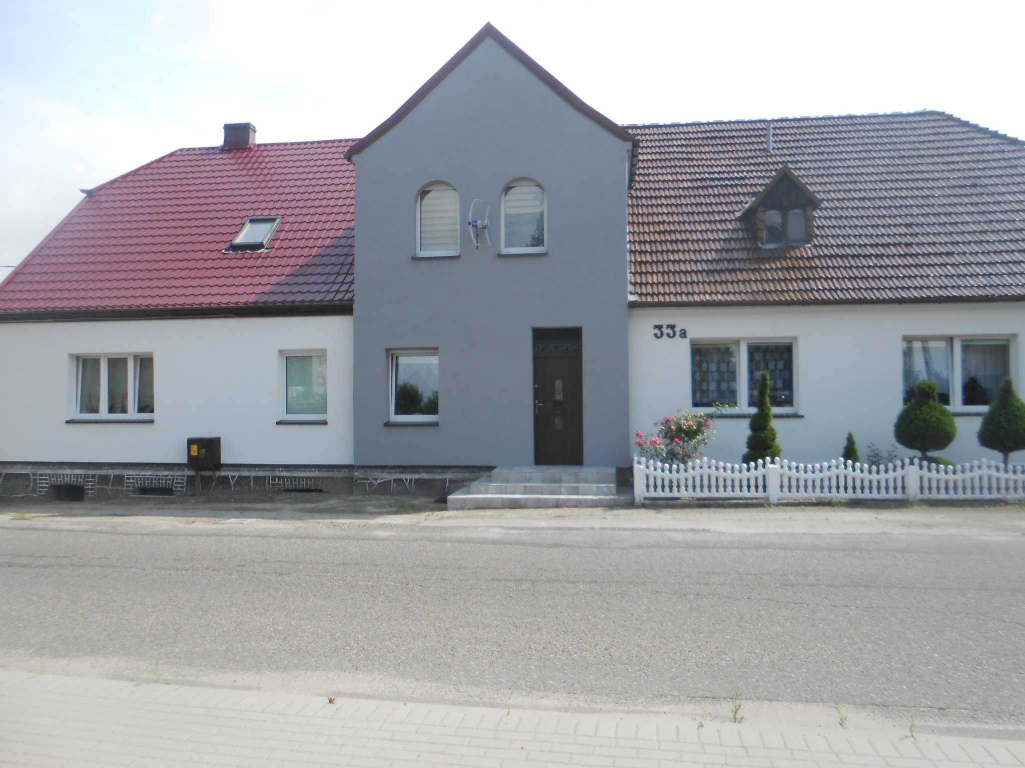 Dom jednorodzinny po renowacji elewacji z widoczną nową szarą częścią środkową, czerwonym dachem po lewej stronie i brązowym po prawej stronie, białe ściany boczne i białe ogrodzenie.