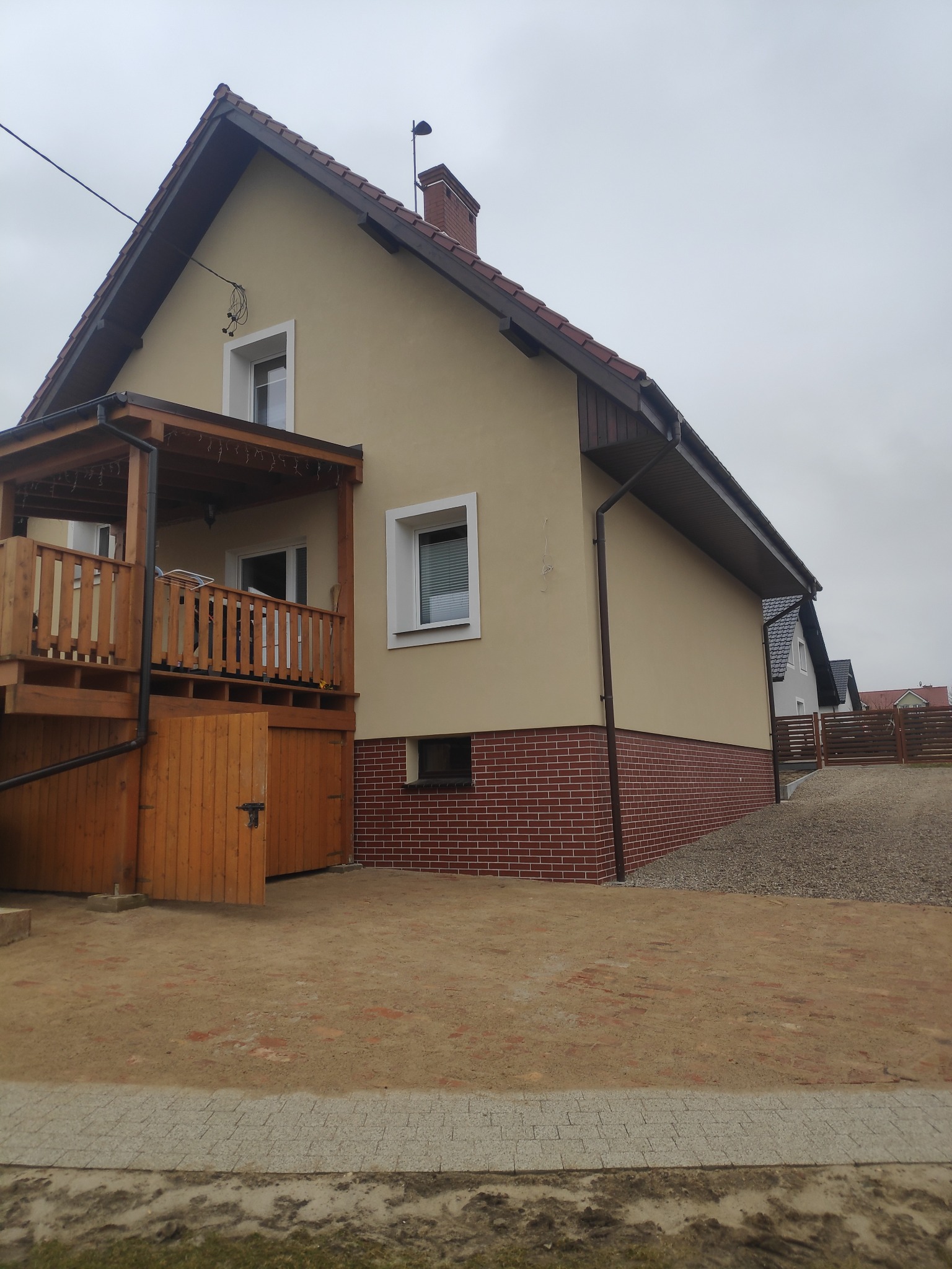 Jasnożółty dom jednorodzinny z drewnianym balkonem i ceglaną podmurówką, widoczny z boku, na tle pochmurnego nieba.