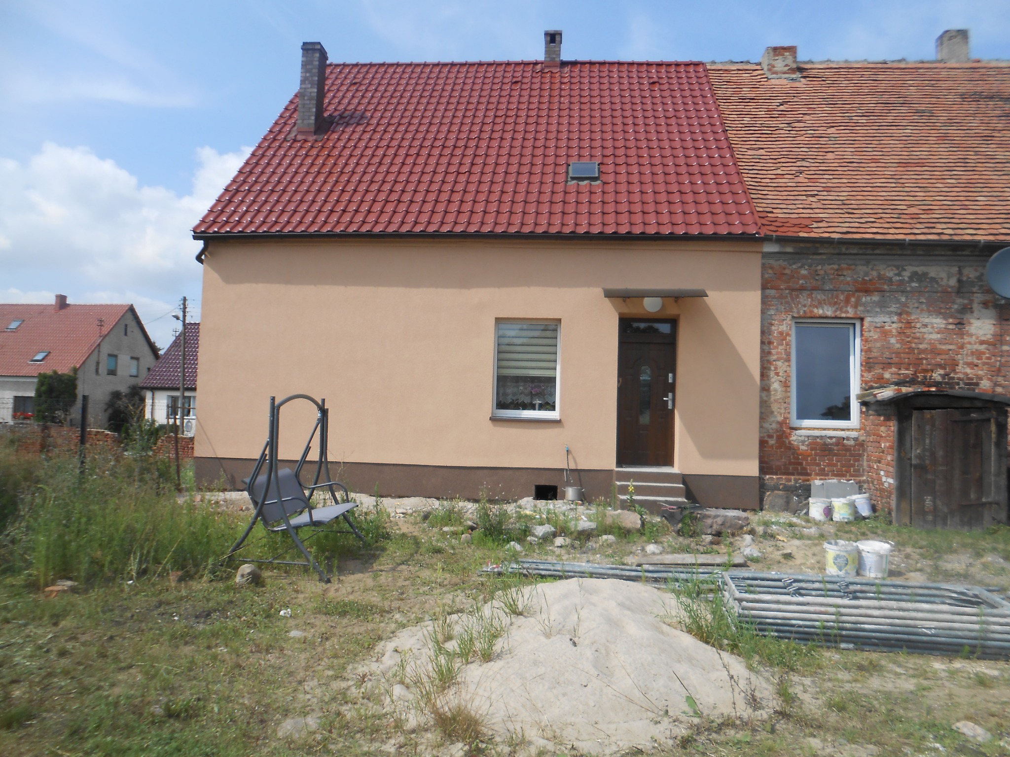 Odnowiony dom z nową, czerwoną dachówką i beżową elewacją, kontrastujący z sąsiednią, starszą częścią budynku z cegły i starą dachówką. Na pierwszym planie huśtawka ogrodowa, sterta piasku i rury.
