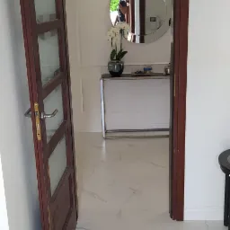 Otwarta, drewniana, przeszklona drzwi wewnętrzne, widok na przedpokój z marmurową podłogą, konsolą z kwiatami i okrągłym lustrem.