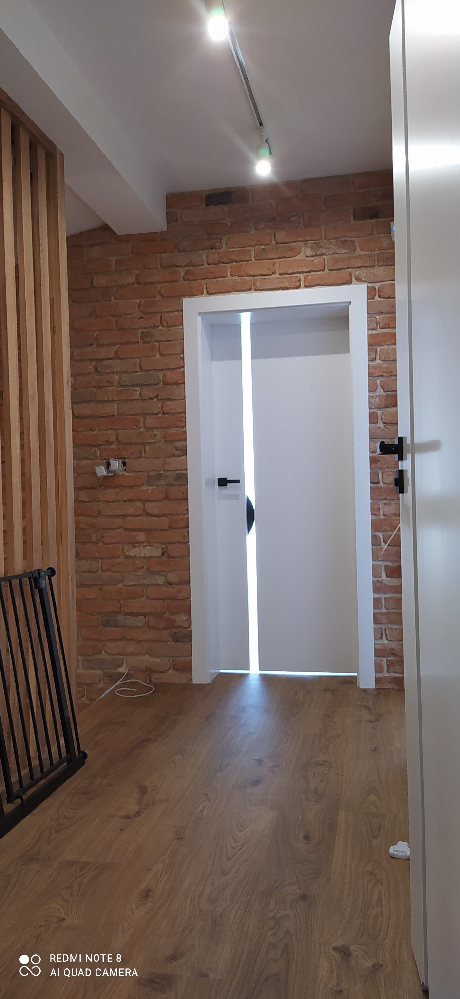 Wnętrze z ceglaną ścianą, drewnianą podłogą i białymi drzwiami z czarnymi klamkami, oświetlone punktowymi lampami sufitowymi.