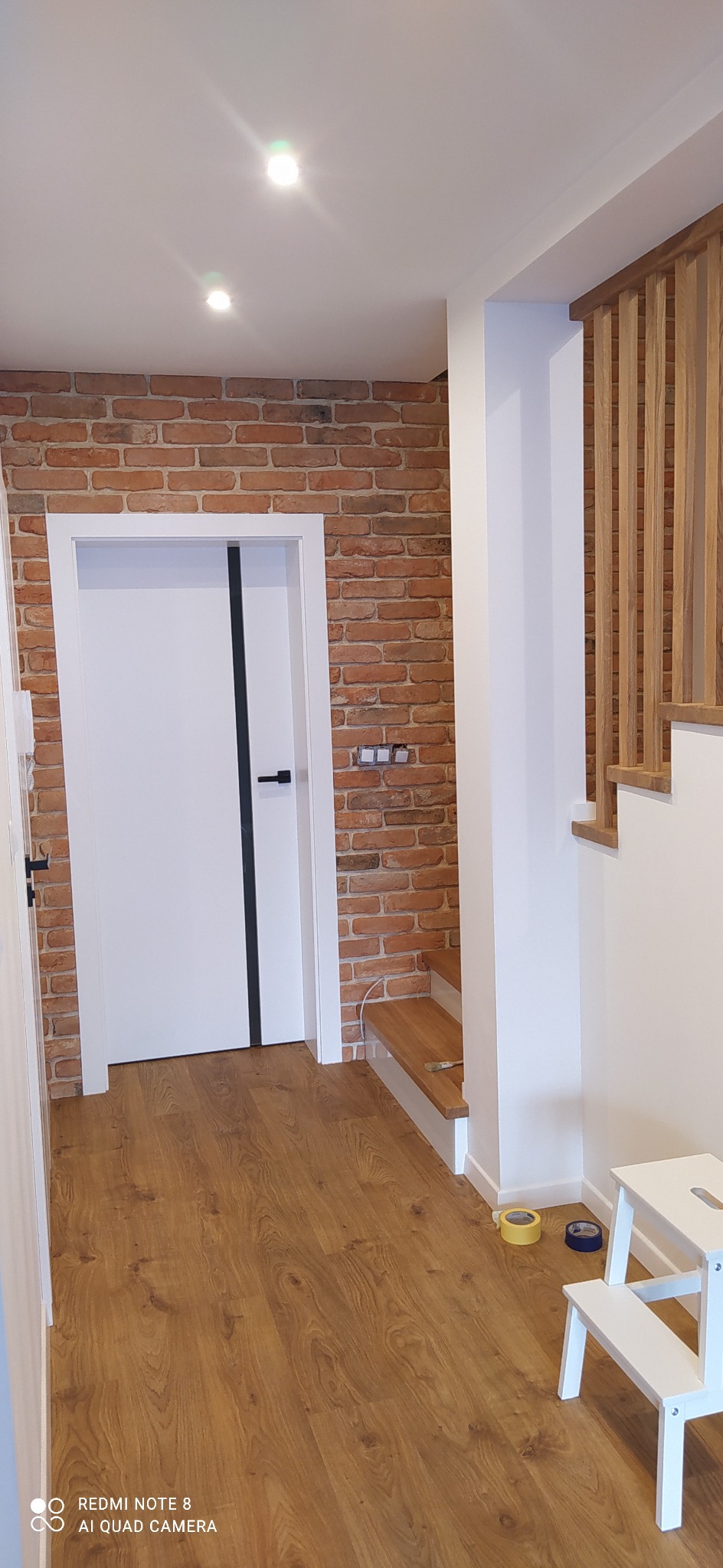 Korytarz po remoncie z ceglaną ścianą, białą stolarką drzwiową z czarnym akcentem, drewnianymi schodami i panelami podłogowymi, widoczne taśmy malarskie i taboret malarski.