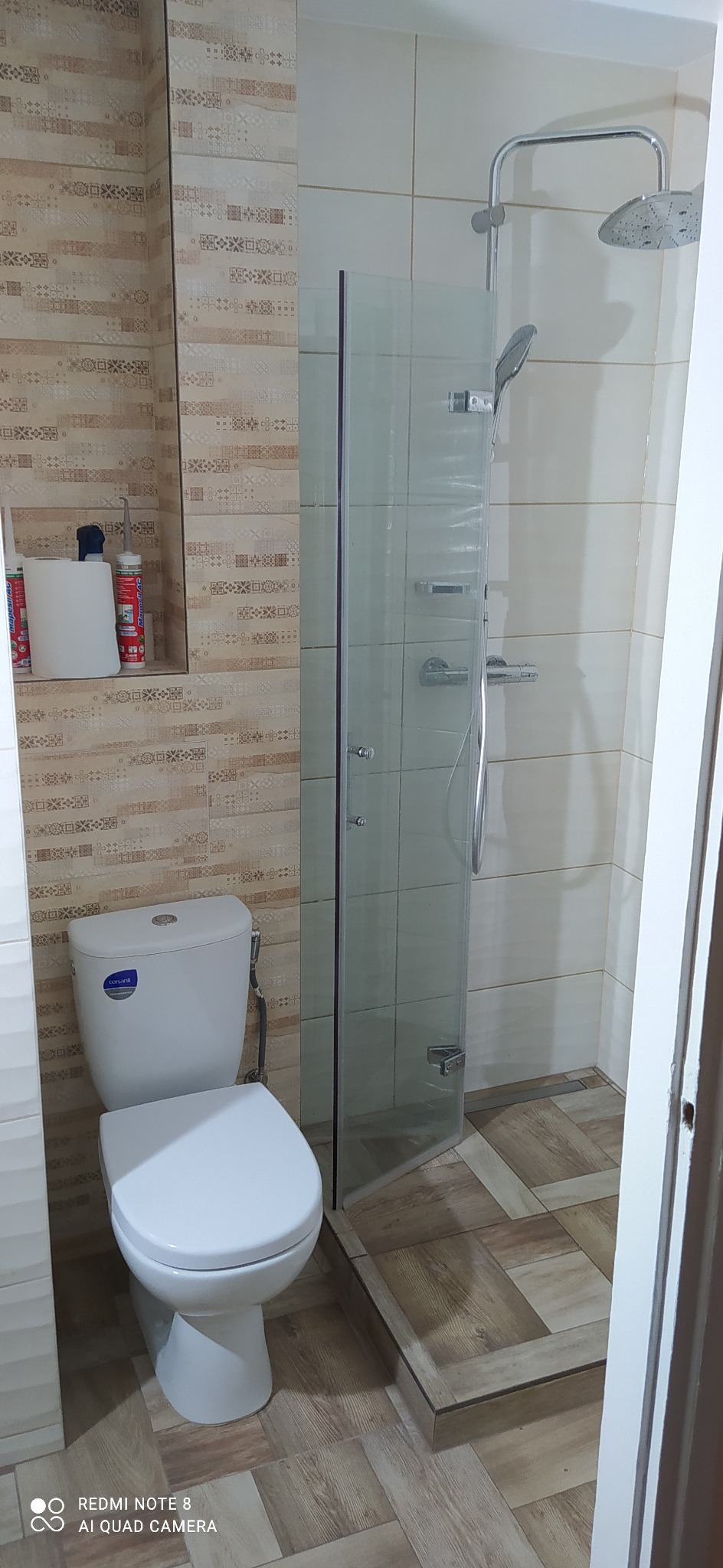 Nowoczesna łazienka po remoncie z kabiną prysznicową ze szklanymi drzwiami, toaletą Cersanit i beżowymi płytkami imitującymi drewno na ścianach i podłodze, z widocznymi silikonami sanitarnymi w półce.