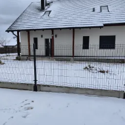 Nowoczesny dom jednorodzinny z białymi ścianami i ciemną stolarką okienną, otoczony metalowym ogrodzeniem panelowym w zimowej scenerii, z widocznymi śladami na śniegu.