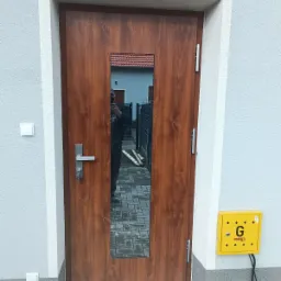 Brązowe drzwi wejściowe z przeszkleniem w Skarbimierz Osiedle, widoczna skrzynka gazowa na ścianie i odbicie fotografa w szybie.