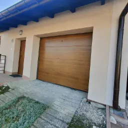 Zewnętrzna, segmentowa brama garażowa w kolorze jasnego drewna, zamontowana w beżowej elewacji budynku z numerem 46, widoczna z poziomu chodnika wyłożonego płytami.