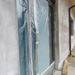 Montaż okna PCV w surowym stanie, zabezpieczonego folią ochronną, widoczne niedokończone ocieplenie i surowy beton.